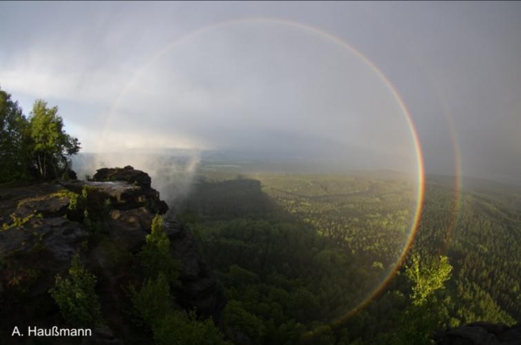 3/4ths of a circular rainbow and its secondary over a lush forest. One quarter of the rainbow is obscured by a part of the mountain.