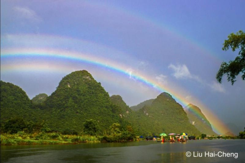 A multi-split rainbow over a body of water and mountains. At the center is a bright primary rainbow. Just below it is a fainter slightly wider rainbow. Right bove it is a fainter rainbow, its outer red edge just visible, converging into the red of the primary near the horizon.