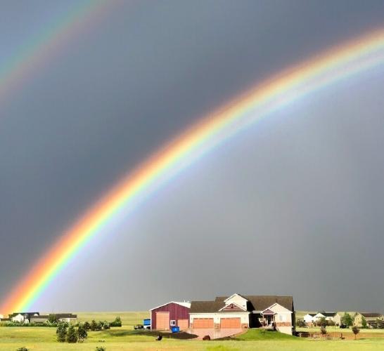 rainbow over a house against a dark sky. The arc starts normal at the horizon, but loses its green and blue further up, instead fading into white, a dark band, and then white again.